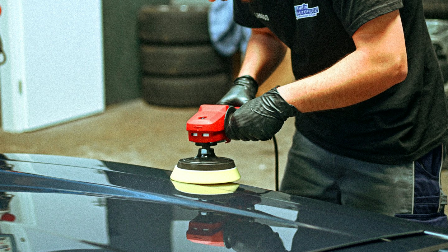 Man met handschoenen polijst motorkap van auto met polijstmachine.